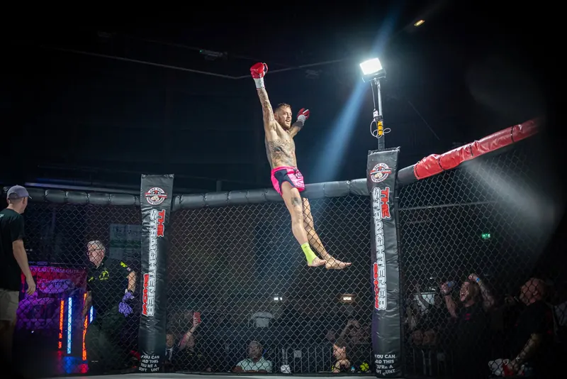 Winning fighter leaping on the cage with his arms raised under a spotlight