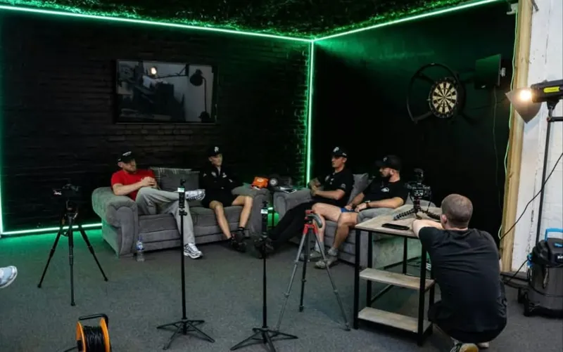 Four guests on grey sofas recording a podcast on the green LED lounge set, with cameras on tripods and a studio light
