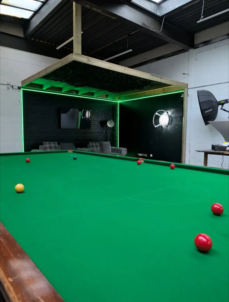 Close view across the green baize of the studio snooker table towards the LED lounge set with a dartboard and studio softbox